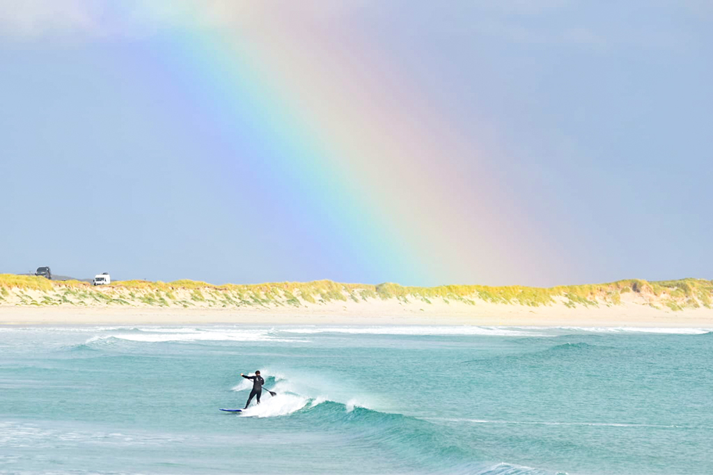 A stand-up paddleboarder catches a turquoise wave close to a sandy beach, set against a bright sky featuring a vibrant rainbow. The surfer, clad in a dark wetsuit, balances on a blue board while carving through a small breaking wave.